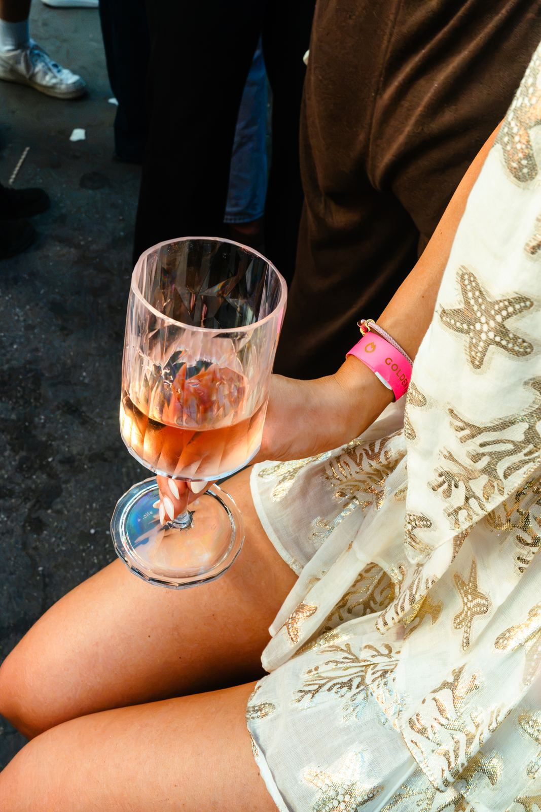 Hand hält ein Glas Rosé über einem Sommerkleid mit goldenen Sternen, pinkes Event-Bändchen am Handgelenk